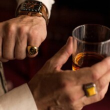 Man looking at his watch on the left hand with a ring on the little finger. In right hand he holding a glass of whiskey.